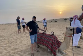 wielbłąd siedzący na pisku pustyni obok turysta